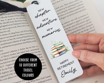 May include: A white rectangular bookmark with a black tassel, featuring the text "NEW chapter NEW adventure NEW memories" and "HAPPY RETIREMENT Emily" in black script and print, along with a watercolor illustration of a stack of four books.