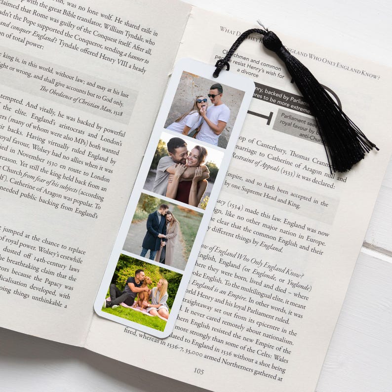 May include: A rectangular white bookmark featuring four vertical photo slots displaying various couples in outdoor settings. The bookmark is topped with a black fabric tassel and is positioned resting on the open pages of a printed book.
