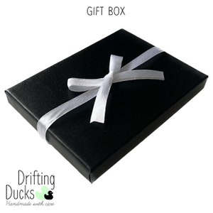 Pu&ograve; includere: Una scatola regalo nera legata con un nastro di raso bianco. Il testo "GIFT BOX" &egrave; in alto e il logo "Drifting Ducks Handmade with care" &egrave; in basso.