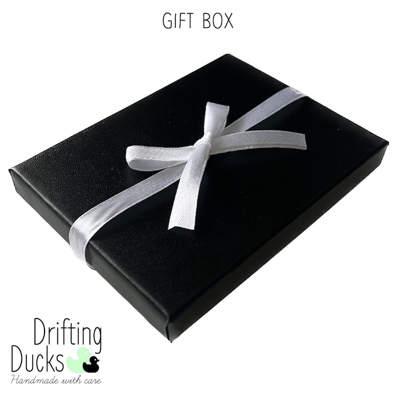 Pu&ograve; includere: Una scatola regalo nera legata con un nastro di raso bianco. Il testo "GIFT BOX" &egrave; in alto e il logo "Drifting Ducks Handmade with care" &egrave; in basso.