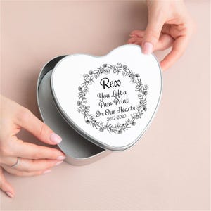 May include: Heart-shaped, silver-colored tin with a white lid. The lid features a black floral wreath design around the text "Rex You Left a Paw Print On Our Hearts 2012-2020". The tin is open, revealing a gray interior.
