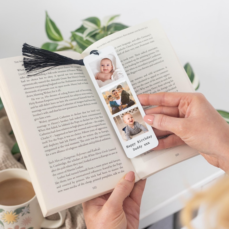 May include: A personalised bookmark with three photo slots and a black tassel. The bookmark features three family photos and the text "Happy Birthday Daddy xxx". The bookmark is held open in a book.