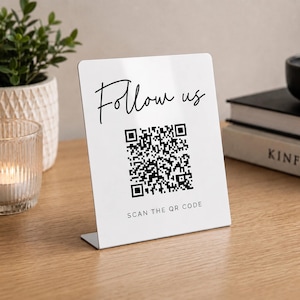 May include: A white rectangular acrylic sign with a bent base for standing. The sign features the black text Follow us in a script font, a central black and white QR code, and the text SCAN THE QR CODE in a simple sans-serif font below it.