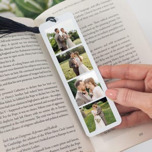 May include: A white rectangular bookmark featuring four vertical photo panels of a couple in a field. The bookmark has a black tassel attached to the top and is placed inside an open book with printed text on the pages, showing a couple in various poses.