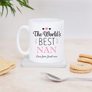 Peut inclure: Un mug en céramique blanche avec le texte noir The World's BEST NAN, une icône de cœur rose au-dessus du mot BEST et le texte NAN en rose. En dessous, le texte Love from Jacob xxx est imprimé en écriture noire. La tasse a une anse à gauche.