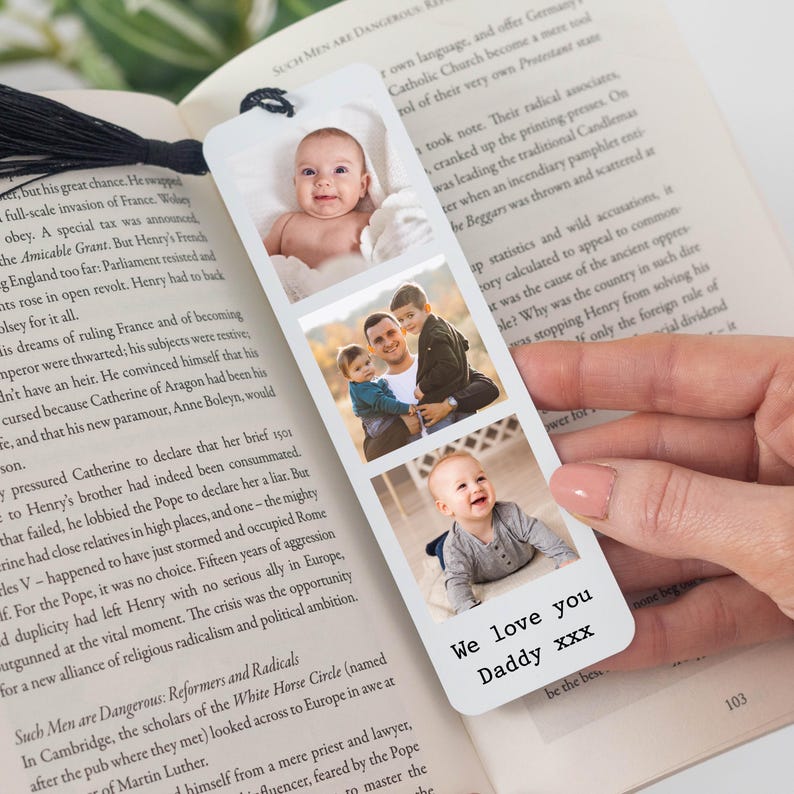 May include: A white bookmark with three family photos and the text "We love you Daddy xxx". The bookmark is held over an open book. The bookmark is a sentimental gift.