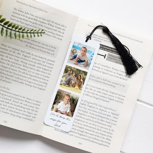 May include: A personalized bookmark with three family photos and a black tassel. The bookmark has the text "We'll keep the page for you Dad". It is placed in an open book.