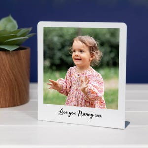 May include: A white polaroid-style photo frame featuring a smiling toddler in a floral dress. The photo includes the text "Love you Nanny xxx". The background is a blurred outdoor scene.