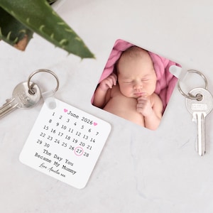 May include: A square keychain with a baby photo and a calendar for June 2026, with the 27th circled. Text reads "The Day You Became My Mummy." The keychain is attached to a keyring with keys.