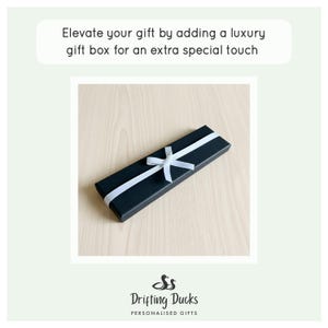 May include: A rectangular black gift box tied with a white ribbon. The text above the box reads: Elevate your gift by adding a luxury gift box for an extra special touch. Below the box is the logo for Drifting Ducks Personalised Gifts.
