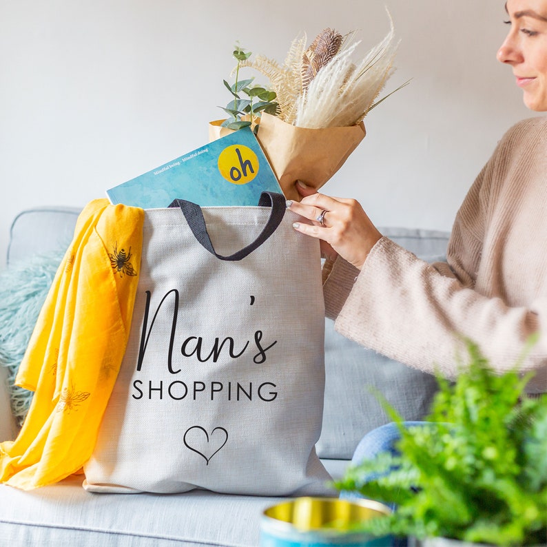 May include: A white canvas tote bag with black handles and the text "Nan's Shopping" printed in black. A small heart is printed below the text. The bag is filled with a bouquet of dried flowers and a magazine.