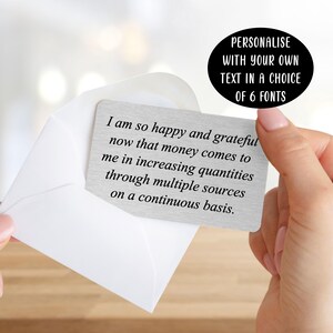 May include: A rectangular silver-toned metal card featuring black engraved text that reads: I am so happy and grateful now that money comes to me in increasing quantities through multiple sources on a continuous basis. The card is tucked into a white envelope.