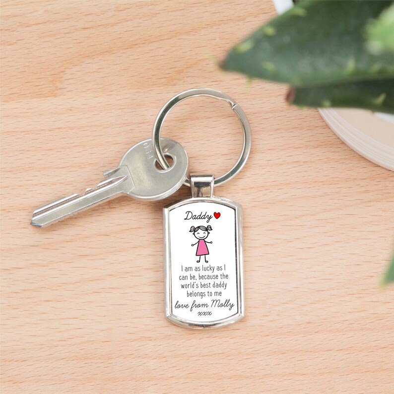 A close-up shot of a silver rectangular metal keyring attached to a single silver key. The keyring features a white insert with "Daddy and a love heart" at the top, a simple stick figure drawing of a girl in a pink dress, and the text "I am as lucky as I can be, because the world's best daddy belongs to me. love from Molly xxxx". The keyring and key are lying on a light wooden surface with a blurred green plant in the background on the right.