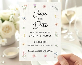 May include: A white rectangular Save the Date card with a subtle grid texture, featuring delicate watercolor floral illustrations along the borders. The text reads: Save THE Date FOR THE WEDDING OF LAURA & JAMES 24.07.2027 ISCOYD PARK, WHITCHURCH formal invitation to follow.