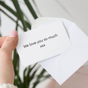 Pu&ograve; includere: Un biglietto bianco con angoli arrotondati all'interno di una busta bianca. Il biglietto recita "We love you so much" in testo nero, seguito da "xxx". Lo sfondo include una pianta verde e un colore neutro sfocato.