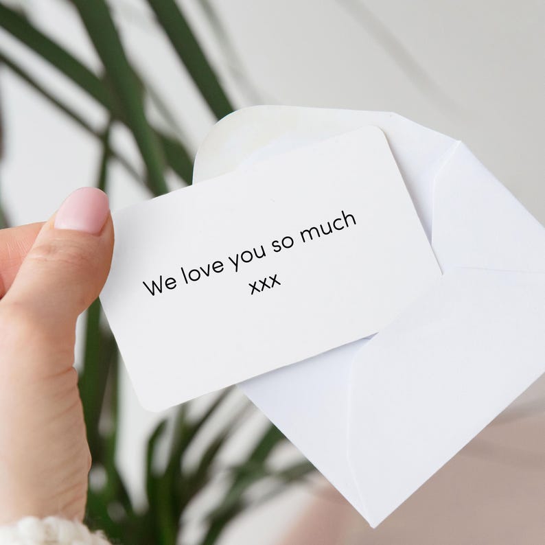 Pu&ograve; includere: Un biglietto bianco con angoli arrotondati all'interno di una busta bianca. Il biglietto recita "We love you so much" in testo nero, seguito da "xxx". Lo sfondo include una pianta verde e un colore neutro sfocato.