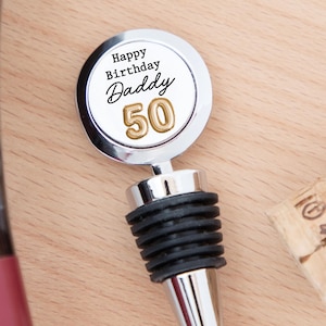 May include: A silver-toned metal bottle stopper featuring a circular top with a white insert. The text reads Happy Birthday Daddy in black, with the number 50 in gold below it. The stopper has a black ribbed rubber seal for a secure fit in bottles.