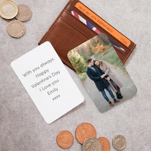 Pu&ograve; includere: Un portafoglio in pelle marrone con una fototessera e una tessera bianca con testo. La fototessera mostra una coppia. La tessera bianca dice "With you always. Happy Valentine's Day I Love you Emily xxxx". Monete sparse.