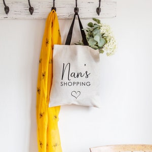 May include: A white tote bag with black handles and the text "Nan's Shopping" with a small heart underneath. The bag is hanging on hooks with a yellow scarf draped over the hooks.