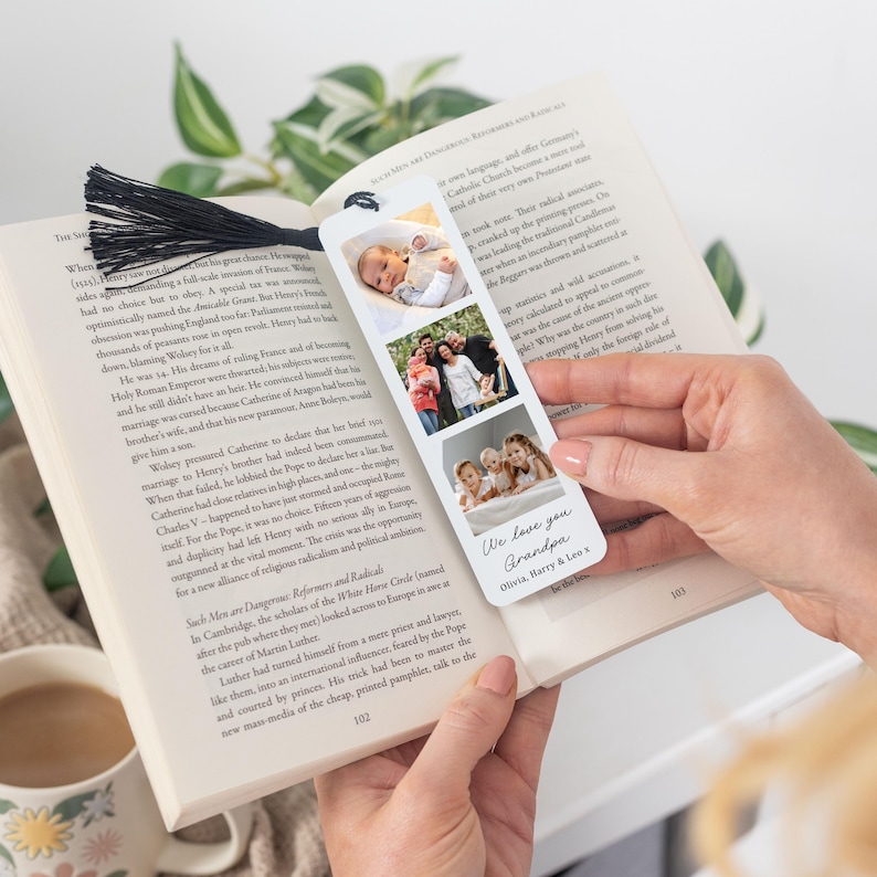 May include: A personalized bookmark featuring three family photos and the text "We love you Grandpa." The bookmark has a black tassel and is held open in a book. A mug is visible in the lower left corner.