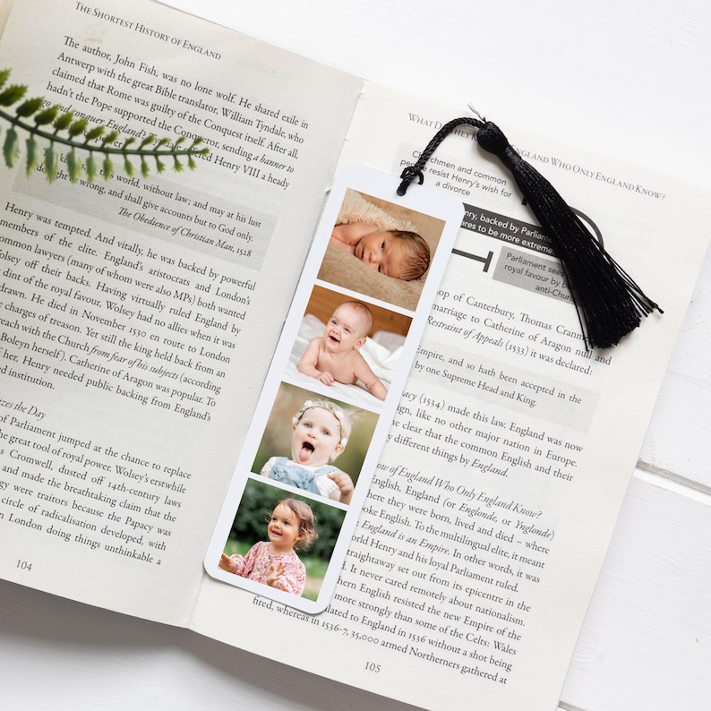 May include: A white rectangular bookmark featuring four vertical photographic portraits of a child at different ages, ranging from infancy to toddlerhood. The bookmark is topped with a black fabric tassel and is placed inside an open book.