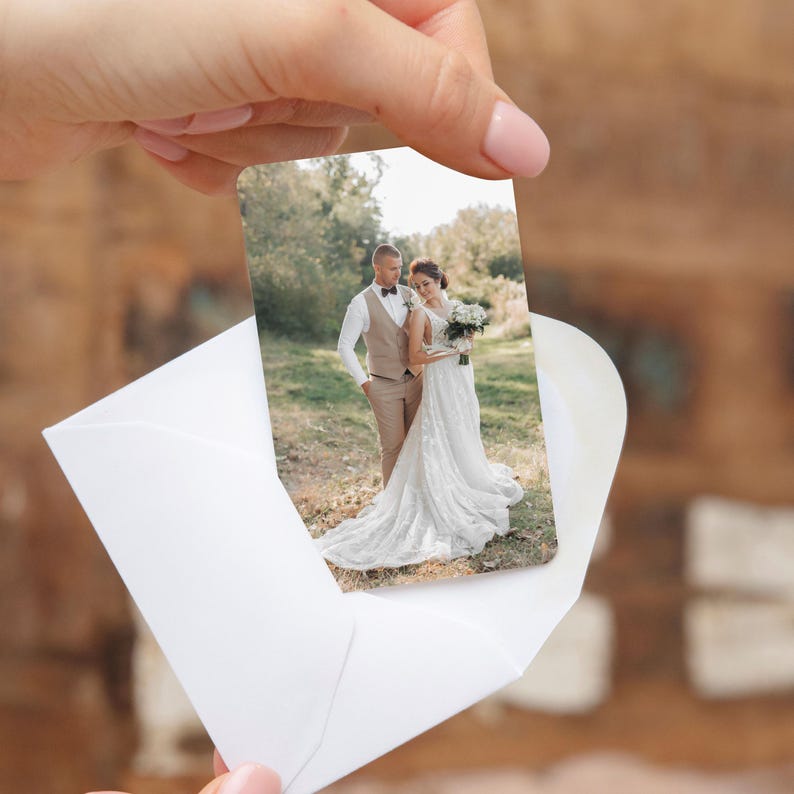 Pu&ograve; includere: Una cartolina fotografica di matrimonio con una sposa e uno sposo in abito formale, tenuta in una busta bianca. La cartolina mostra la coppia in piedi all'aperto, la sposa in un abito bianco e lo sposo in un abito. La cartolina &egrave; tenuta da una mano.