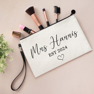 May include: A cream-colored canvas zippered pouch with a black wrist strap. The front features the black text Mrs Harris, EST 2024, and a small heart outline. Inside the pouch are three makeup brushes and a pink lipstick with a gold cap.