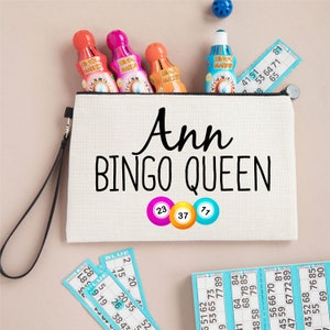 May include: A white canvas zippered pouch with a black handle. The pouch has the text "Ann BINGO QUEEN" printed on it in black. There are three bingo balls with the numbers 23, 37, and 77 printed on them.