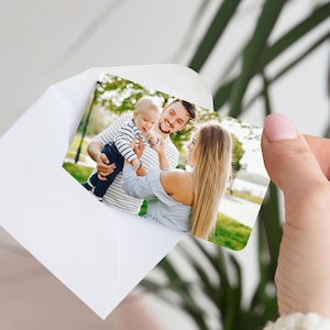 Pu&ograve; includere: Una busta bianca che contiene una fotografia di famiglia. La foto mostra un bambino sorridente tenuto in braccio da un uomo, con una donna che si protende verso il bambino. Lo sfondo &egrave; un parco con alberi.