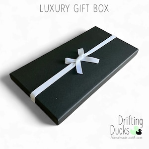 Puede incluir: Una caja de regalo rectangular negra con una cinta y un lazo blancos. El texto "LUXURY GIFT BOX" est&aacute; en la parte superior. El logotipo de "Drifting Ducks Handmade with care" est&aacute; en la esquina inferior derecha.