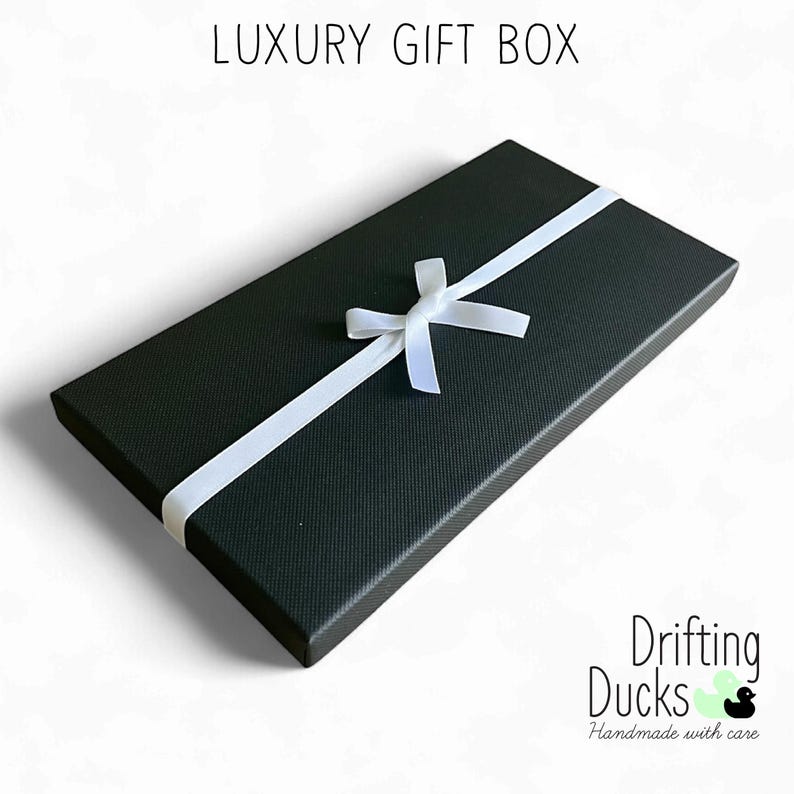 Puede incluir: Una caja de regalo rectangular negra con una cinta y un lazo blancos. El texto "LUXURY GIFT BOX" est&aacute; en la parte superior. El logotipo de "Drifting Ducks Handmade with care" est&aacute; en la esquina inferior derecha.