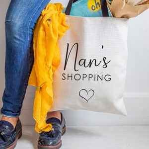 May include: A white canvas tote bag with the text "Nan's Shopping" and a small heart design. The bag is being held by a person wearing blue jeans and black shoes.