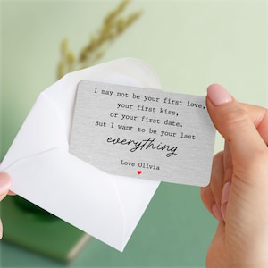 May include: A silver metal card with a romantic message in black font, held in a white envelope. The card reads "I may not be your first love... But I want to be your last everything. Love Olivia" with a small red heart.
