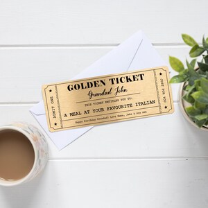 Puede incluir: Un boleto dorado con texto negro que dice "GOLDEN TICKET Grandad John". El boleto est&aacute; sobre un sobre blanco. El texto del boleto dice "A MEAL AT YOUR FAVOURITE ITALIAN". Una taza de caf&eacute; y una planta tambi&eacute;n est&aacute;n en la imagen.