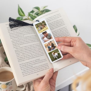 May include: A rectangular white bookmark featuring three vertical photo panels showing a family and a dog, topped with a black tassel. The bookmark is placed inside an open book with printed text on the pages, displaying a clear view of the photos.