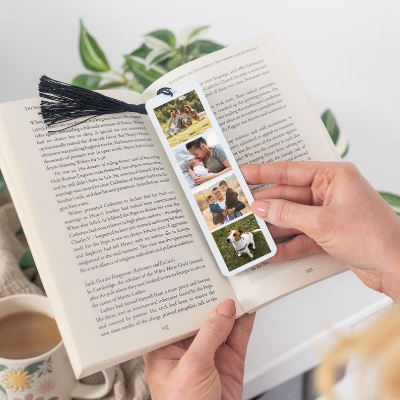 May include: A rectangular white bookmark featuring three vertical photo panels showing a family and a dog, topped with a black tassel. The bookmark is placed inside an open book with printed text on the pages, displaying a clear view of the photos.