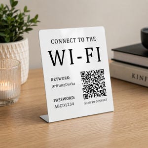 May include: A white rectangular sign with black text that reads CONNECT TO THE WI-FI, NETWORK: DriftingDucks, PASSWORD: ABCD1234, and SCAN TO CONNECT, featuring a black and white QR code on the right side, standing upright on a flat surface.