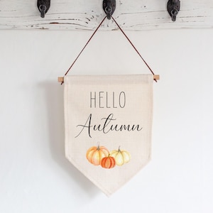 May include: A cream-coloured fabric wall banner featuring the black text HELLO Autumn and a watercolour illustration of three pumpkins in shades of orange and yellow. The banner is suspended from a wooden dowel with a thin brown cord for hanging.