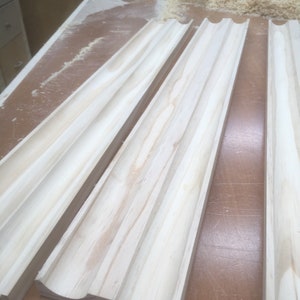 May include: Close-up of several unfinished wooden molding pieces with a curved design. The wood has a light, natural color and visible grain. The pieces are arranged on a brown surface, with wood shavings in the background.