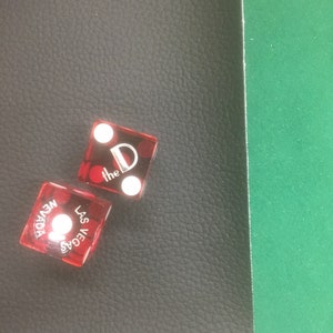 May include: Two transparent red dice with white dots and the text "the D" and "LAS VEGAS NEVADA" are on a black textured surface. One die shows two dots, the other shows one dot. The background is a green surface.