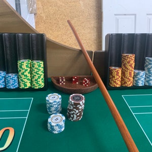 Pode incluir: Uma mesa de jogo de feltro verde com um bastão de madeira, dados vermelhos e pilhas de fichas de pôquer em suportes de fichas pretos, verdes e vermelhos. A mesa tem os números "10" e "4" impressos nela.