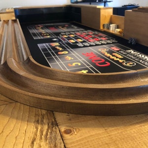 Może przedstawiać: Drewniany stół do gry w kości z czarną filcową powierzchnią do gry. Stół ma zakrzywioną drewnianą krawędź i jest ustawiony na drewnianej powierzchni. Powierzchnia do gry ma nadrukowane obszary do obstawiania, ze słowem "COME" na czerwono.