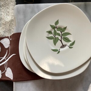 Puede incluir: Una pila de cuatro platos de cerámica blanca con una forma única, ligeramente irregular. Cada plato presenta un diseño central de una planta verde estilizada con hojas. Los platos son parte de un juego de vajilla.