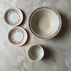 May include: A set of vintage ceramic dishware, including two saucers, a small bowl, and a large bowl. The pieces have a speckled beige background with brown floral patterns and trim. The arrangement is on a neutral surface.