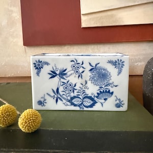May include: A rectangular white ceramic vase with a blue floral design. The vase features a detailed pattern of flowers, leaves, and vines. The design is symmetrical and covers the front and sides of the vase. The vase is sitting on a green surface.