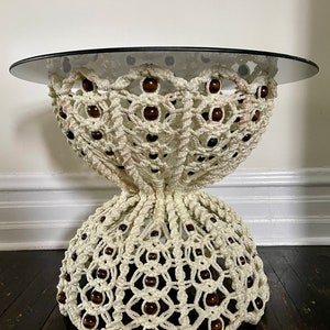 May include: A cream-colored macrame coffee table with a round, dark glass top. The table features a woven design with dark brown wooden beads. The base has a flared shape, and the table is set on a dark wooden floor.