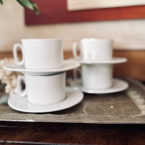 Può includere: Quattro tazze da tè e piattini in ceramica bianca sono impilati su un vassoio d'argento. Le tazze hanno un design semplice con un manico. I piattini sono rotondi e piatti. Il vassoio è rettangolare con bordo decorativo.