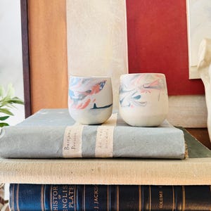 May include: Two small, round ceramic cups with a marbled design in shades of red, blue, and white. The cups are placed on a stack of old books with the titles "History of English Plate" and "Volume I" visible.