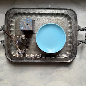 May include: A vintage silver tray with ornate handles holds a small, colorful box, a light blue plate, and dried floral elements. The tray's surface shows signs of wear, adding to its antique charm.