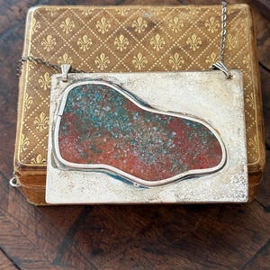 May include: A rectangular silver pendant necklace with a unique design. The pendant features a central, irregularly shaped inlay of red and turquoise stones. The necklace is displayed on a vintage, gold-toned box with a fleur-de-lis pattern.